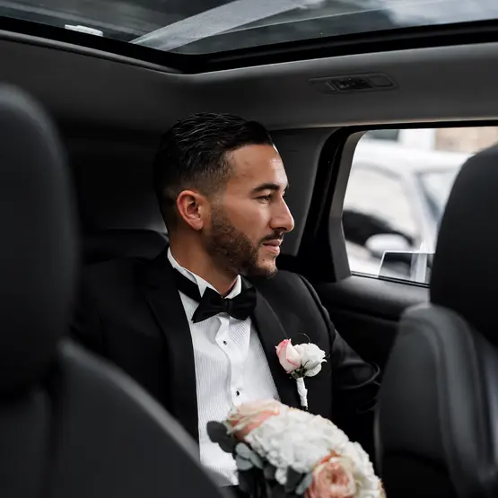 Un marié en costume et nœud papillon, avec un bouquet, assis à l’arrière d’une voiture.