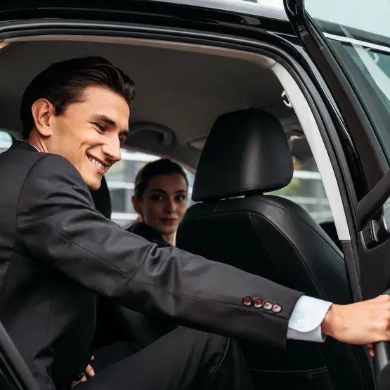 Un passager en costume sourit à bord d’une voiture, avec une autre personne à l’arrière.