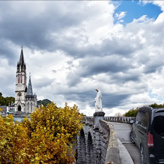 Van stationné près du sanctuaire de Lourdes, avec la basilique et une statue sous un ciel nuageux.